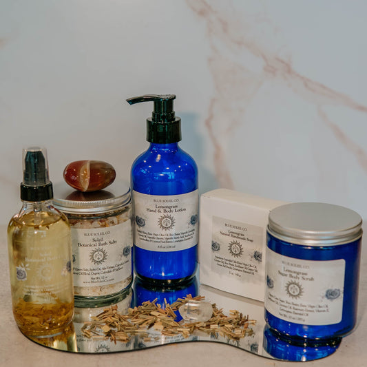 Collection of Blue Soleil Co skincare products including botanical body oil, bath salts, lotion, handmade soap bar, and sugar body scrub bottles and jars on a marble surface.
