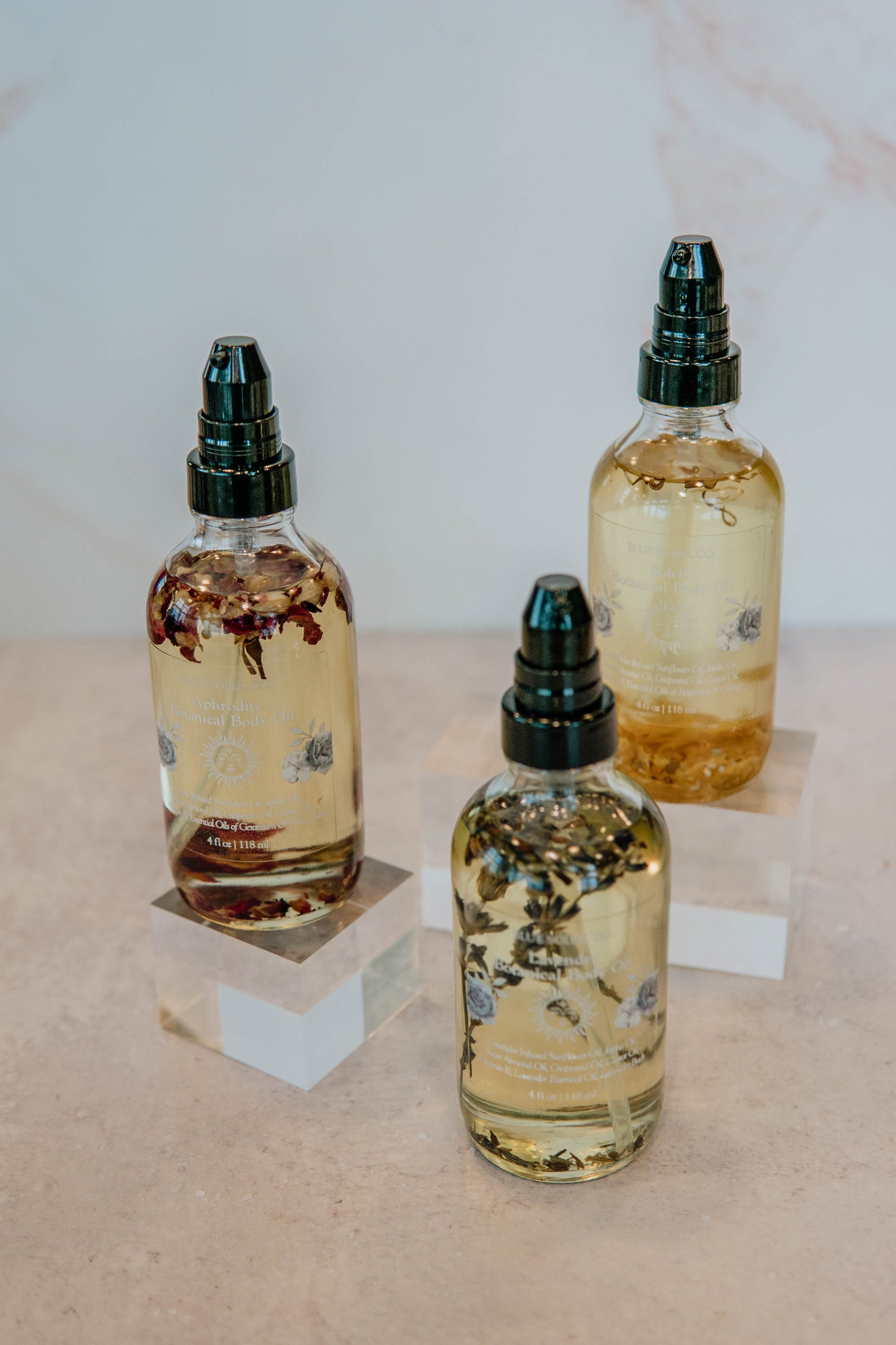 Three 4 oz clear glass bottles with black treatment pump tops with botanical body oil with lavender flowers, roses, and calendula petals. 