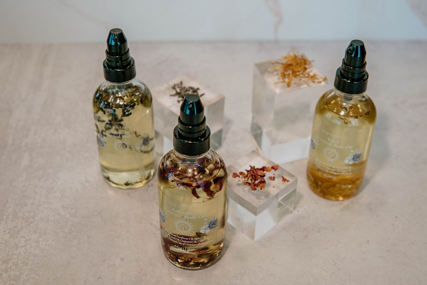 Three 4oz clear glass bottles with black treatment pump tops with botanical body oil with lavender, roses, and calendula flowers.