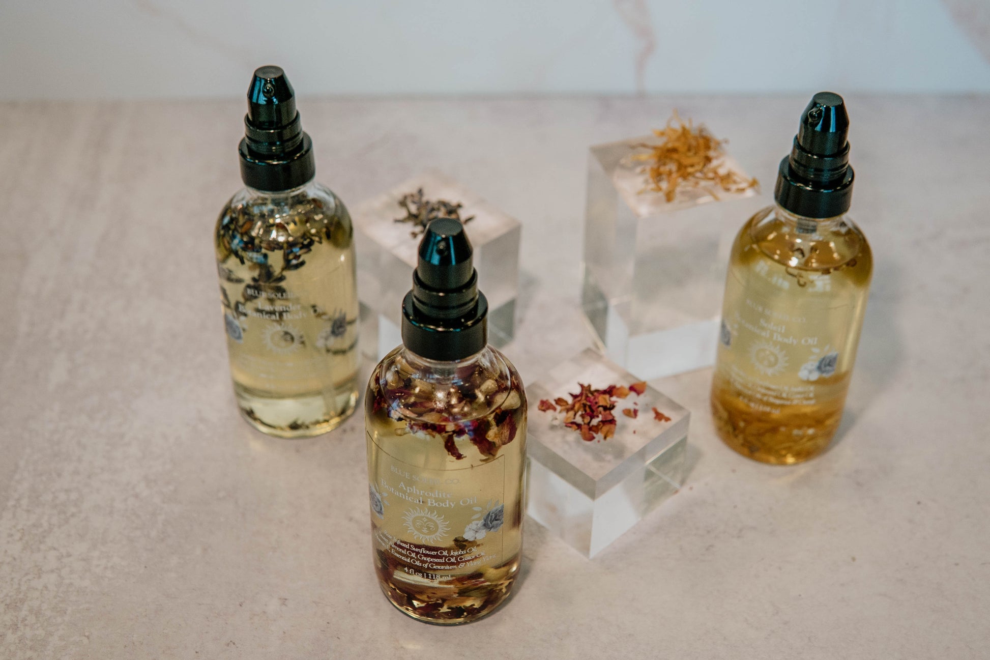 Three 4oz clear glass bottles with black treatment pump tops with botanical body oil with lavender, roses, and calendula flowers.